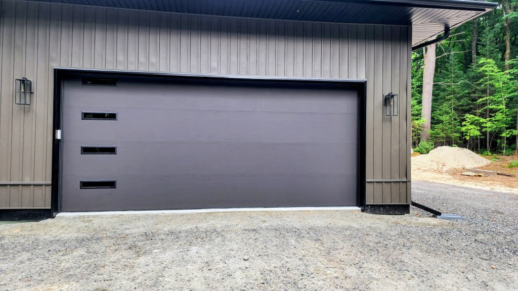 A modern garage door with three horizontal windows, set in a wooden wall of a house, surrounded by gravel and greenery.